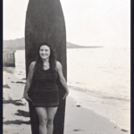 Real Photo Rose Heather stands in front of a surfboard at Gray's Beach by R.J. Baker