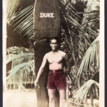 Hand colored Real Photo Duke stands in font of his surfboard at Waikiki Beach, unused