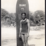 Real Photo Duke stands in front of his board on Waikiki Beach by R.J. Baker, 1912-16