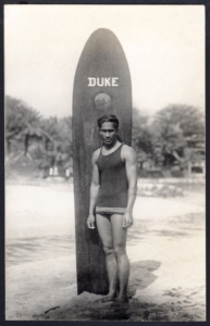 Real Photo Duke stands in front of his board on Waikiki Beach by R.J. Baker, 1912-16